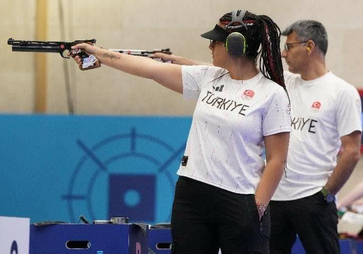 Photo of the two Turkish shooters side by side. They are aiming at a target with similar firearms. Both have one hand in their pocket while shooting. 