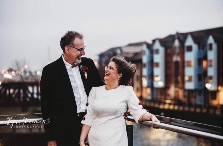 A tall white man in a dark suit and a short white woman in a white dress stand side by side on a balcony, with buildings and a river in the background. The man has his arm around the woman and is smiling at her. She is laughing as her hair is swept back by a strong wind.