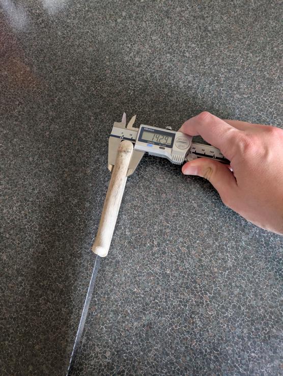 A photo of a bread knife on a kitchen bench, with its handle being measured for width with digital calipers. The readout says 14.29mm.